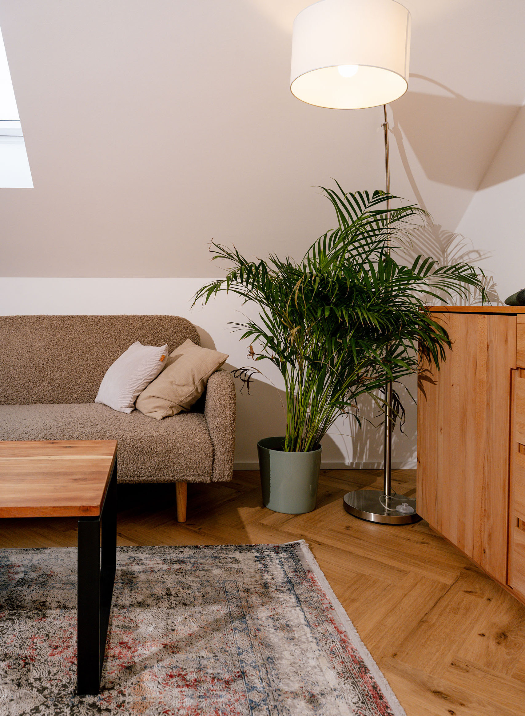 Gemütliche Wohnzimmerecke mit einem hellen Sessel, Kissen, einem Holztisch, einer Stehlampe und einer großen grünen Zimmerpflanze neben einem Holzschrank auf einem gemusterten Teppich.