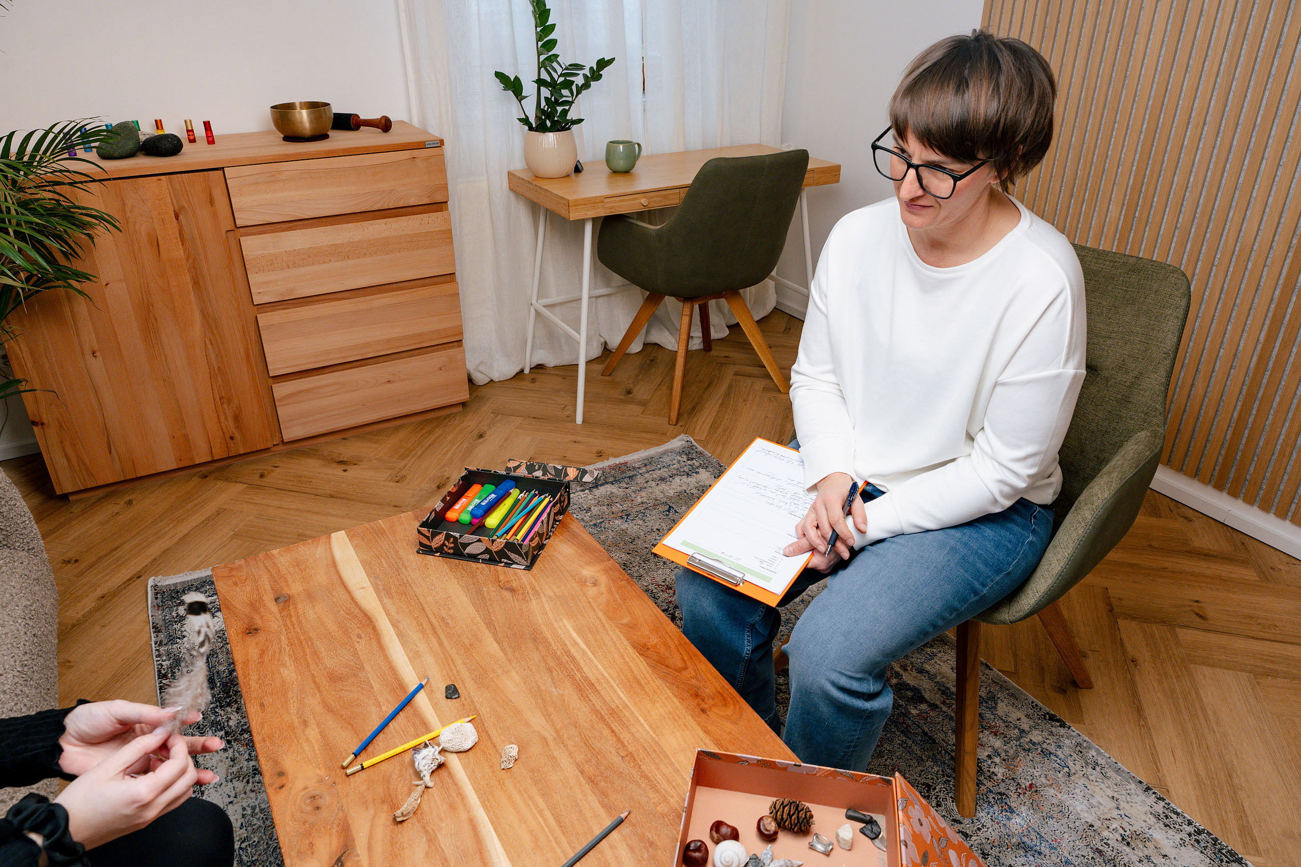Eine Frau sitzt in einem gemütlichen Raum auf einem Stuhl und hält ein Klemmbrett, während sie einer anderen Person gegenüber am Tisch zuhört; auf dem Tisch liegen Stifte, Naturmaterialien und ein offenes Etui.